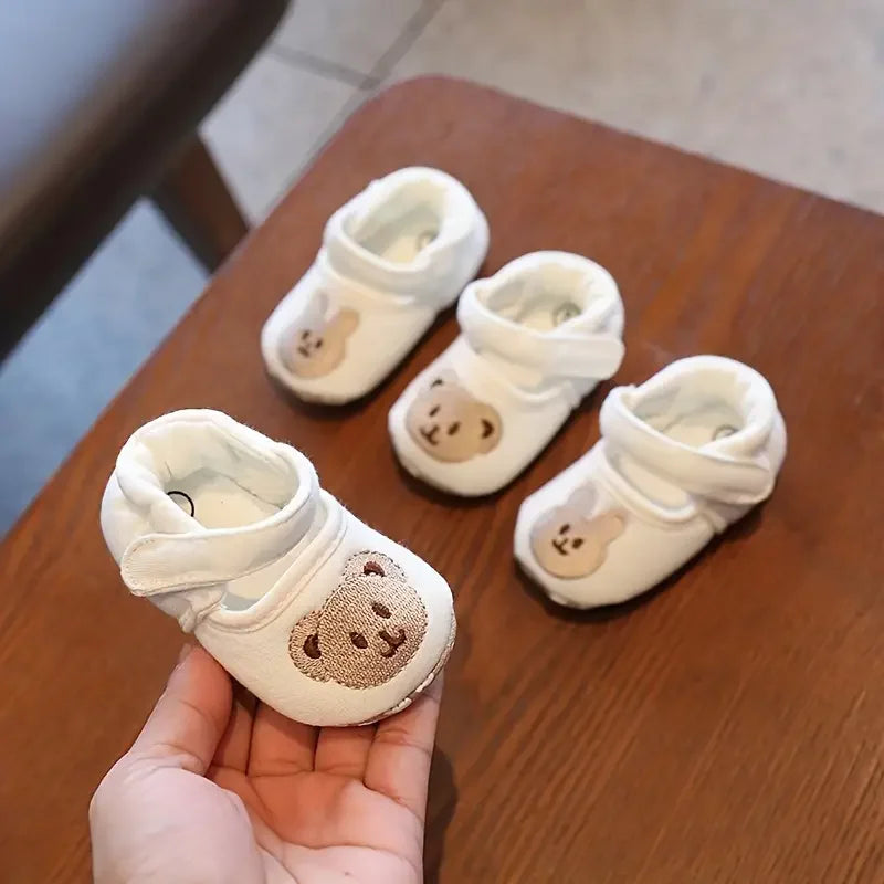 A hand holds a pair of white soft-sole sandals for baby's first steps featuring a cute bear design.