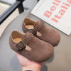 A close-up of a brown suede moccasin shoe with a leather hook-and-loop closure being held in a hand.