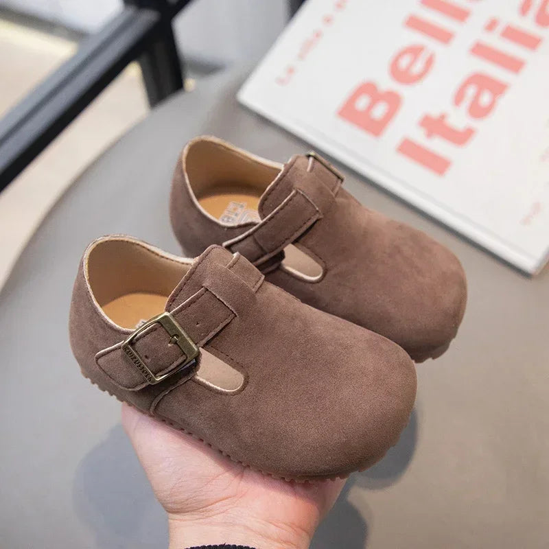 A close-up of a brown suede moccasin shoe with a leather hook-and-loop closure being held in a hand.