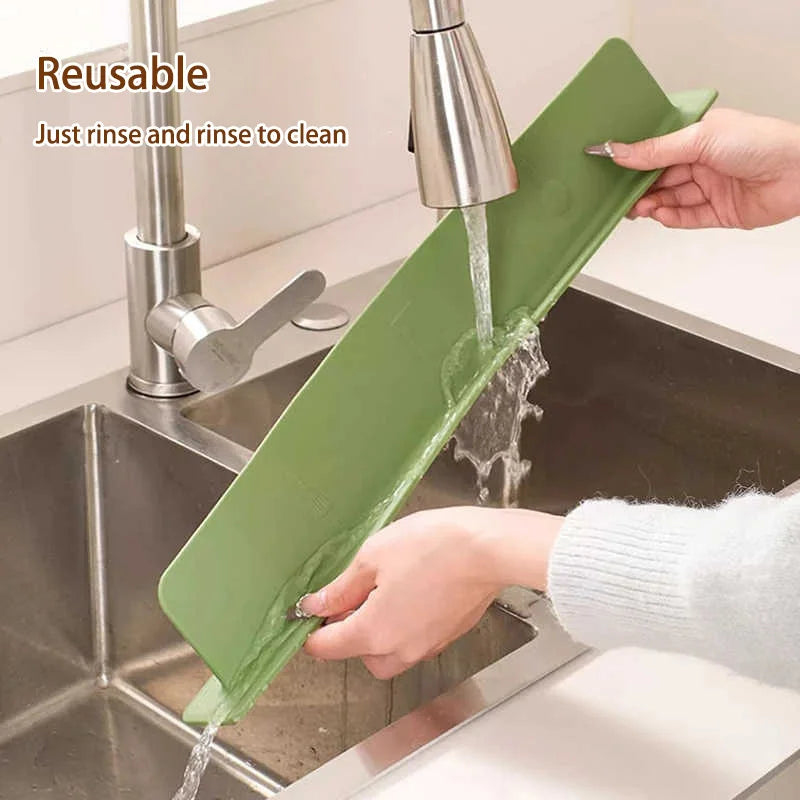 Una mano usa un protector de salpicaduras adhesivo verde para enjuagar y limpiar un fregadero de cocina de acero inoxidable.