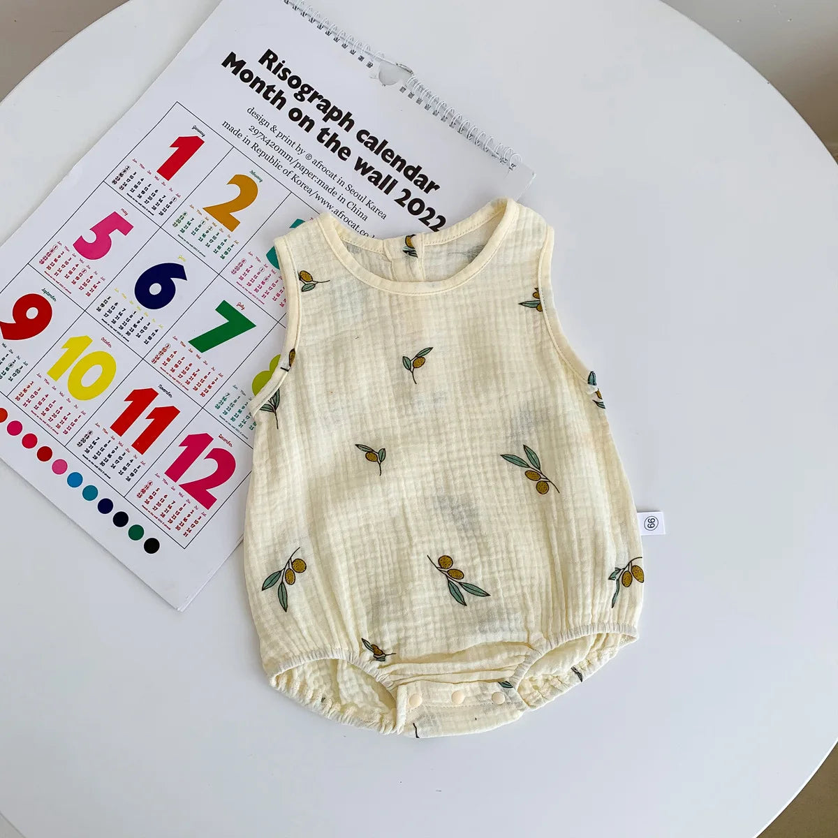 A cream-colored baby cotton sleeveless jumpsuit bodysuit set lying on a white surface next to a calendar.