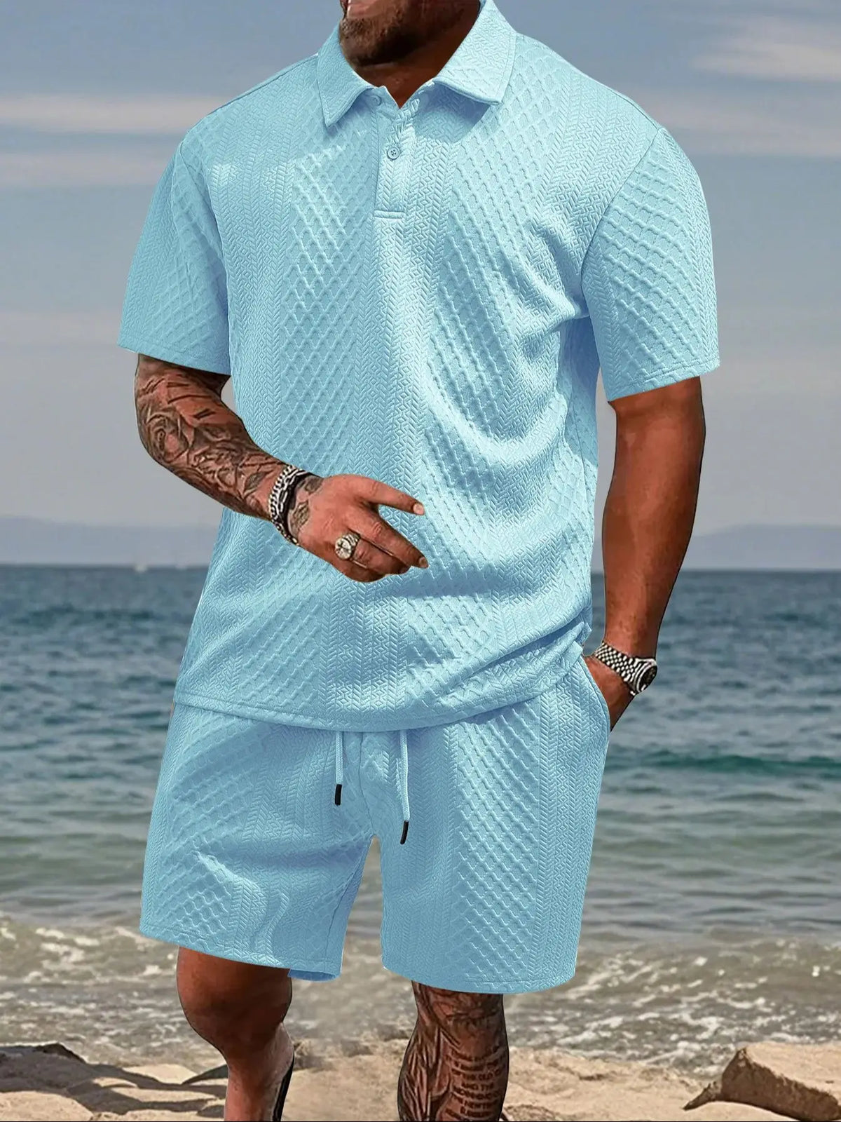 A man wearing a light blue jacquard short-sleeve shirt and matching drawstring shorts outdoors near the water.