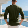 A man wearing a dark olive green long-sleeve shirt and white pants stands outdoors by a pool with a view of the ocean.
