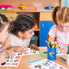 Two young girls are painting on paper with a cupcake diamond painting sticker kit at a wooden table.