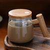 A glass mug filled with light brown liquid is shown on a wooden board, featuring a wooden handle for stirring.