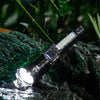 A black rechargeable multi-mode LED flashlight with a telescopic zoom is shown illuminating water droplets on dark rocks.