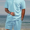 A man wearing a light blue textured polo shirt and matching drawstring shorts standing near the water.