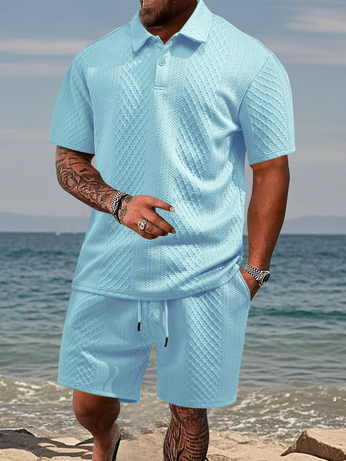 A man wearing a light blue textured polo shirt and matching drawstring shorts standing near the water.