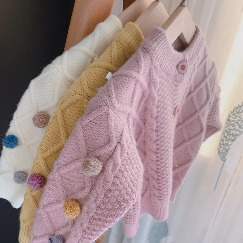 A display of pink knitted cardigan sweaters with various colored pom-pom trims on wooden hangers.
