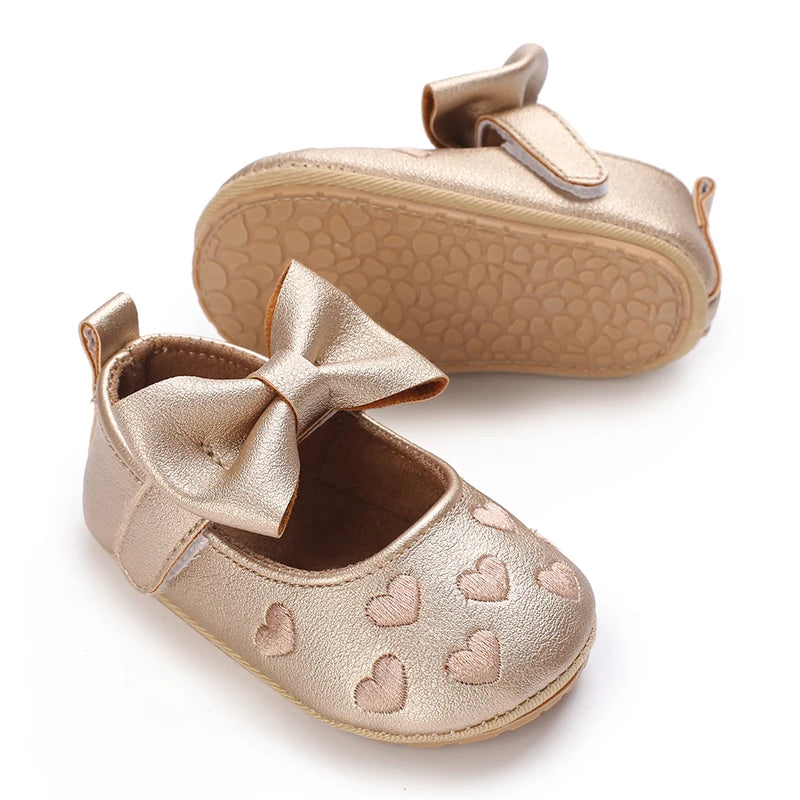 A pair of gold satin baby first walker shoes with a bow and heart patterns.