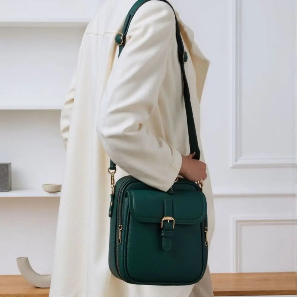 A woman wearing a cream-colored coat is holding a dark green vintage vegan leather messenger bag.
