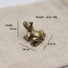 Liten vintage figurin av messingfrosk, 1,5 cm høy og 2,3 cm lang, på lys stoffoverflate.