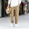 A man wearing tan cargo pants and a white shirt holds a straw hat on a city sidewalk.