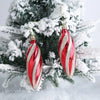 A set of two shiny red and white striped candy cane Christmas tree ornaments hanging on a snowy green fir branch.