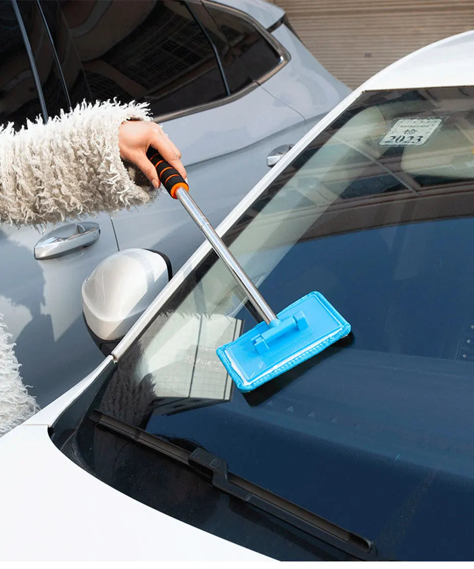 A hand is using a telescopic windshield cleaning brush to wipe the glass of a car.