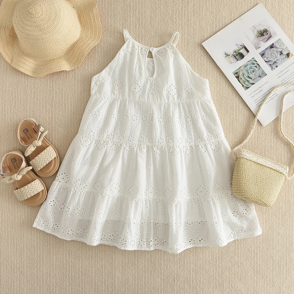 A white cotton dress with eyelet details and smocked bodice, laid out next to a straw hat and sandals.