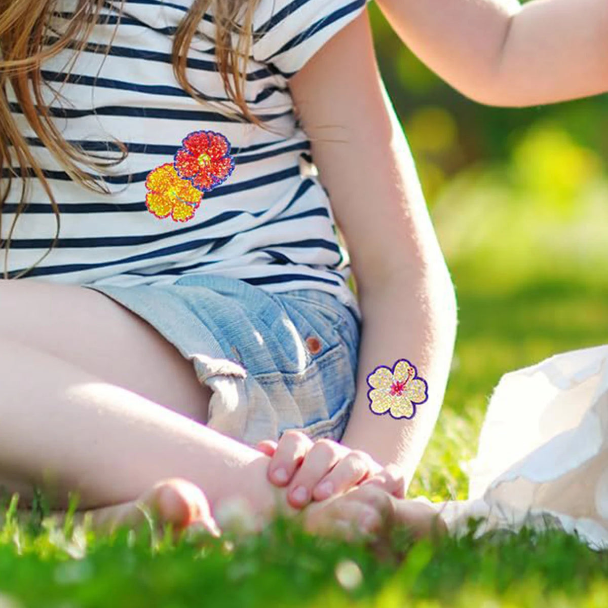 A young child is sitting in the grass with hands clasped, wearing a striped shirt and jeans, near colorful glitter flower sti