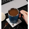 A dark blue stainless steel insulated travel mug sitting on a white coaster next to a laptop and a spoon.