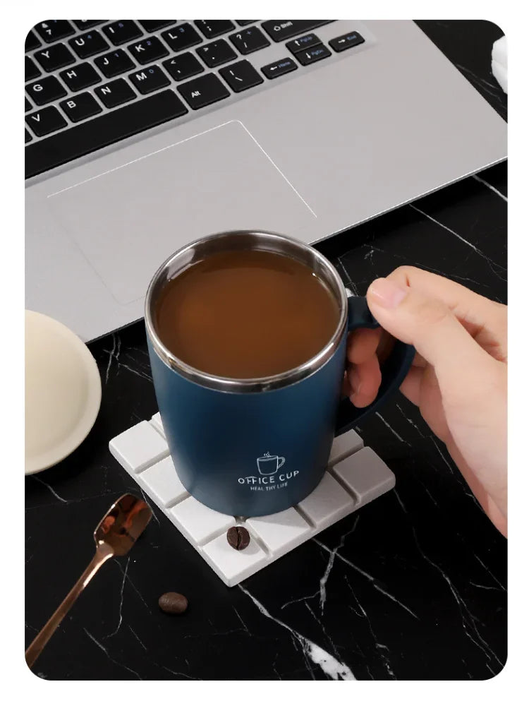 A dark blue stainless steel insulated travel mug sitting on a white coaster next to a laptop and a spoon.