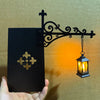 A black metal decorative shelf lamp featuring an ornate scrollwork bracket and a glass lantern with a warm LED glow.