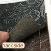 A close-up view of a dark gray textured faux leather fabric with an intricate floral pattern, being held by a finger.