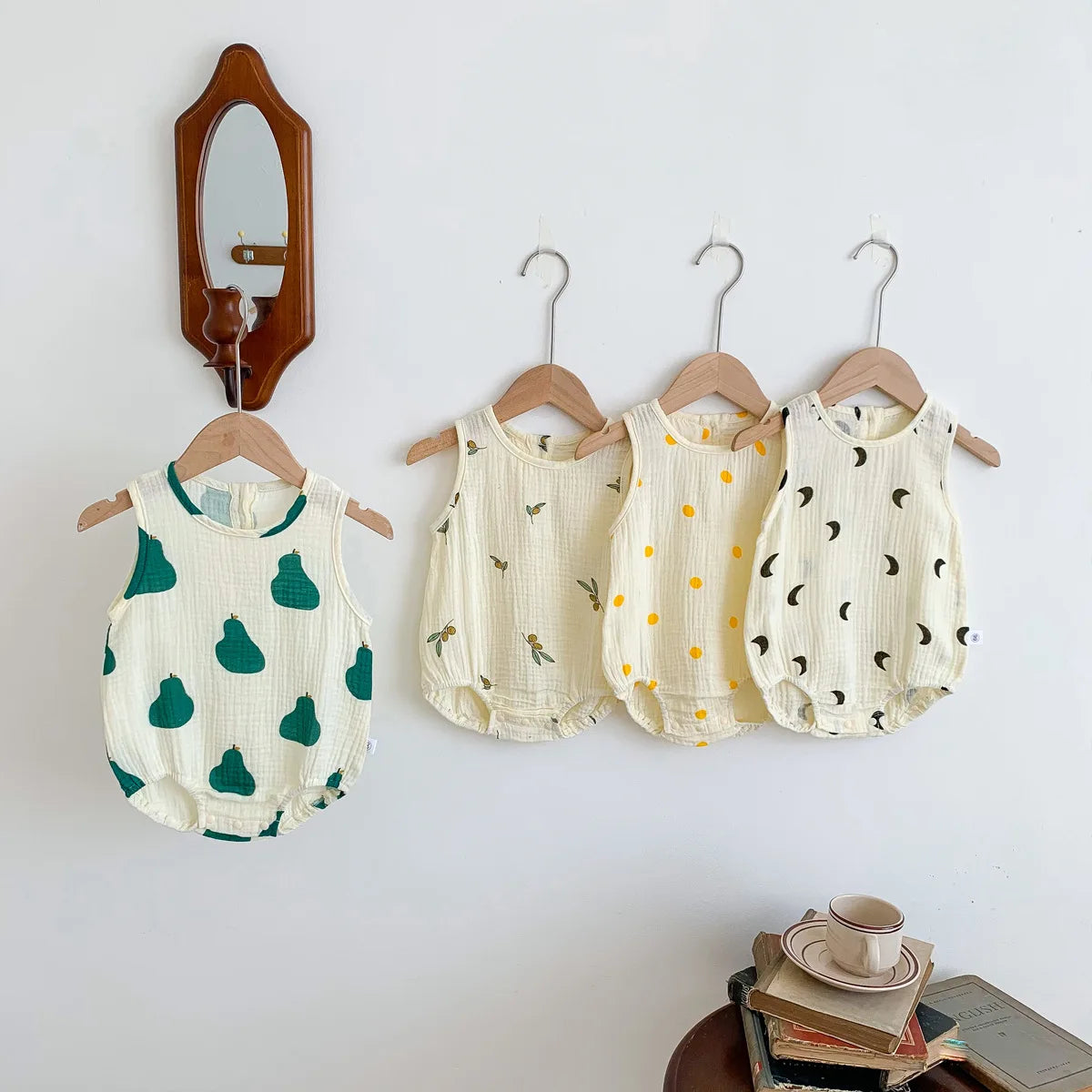 A set of cream and green baby cotton sleeveless jumpsuit bodysuits hanging on a white wall.