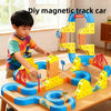 A boy is building a colorful magnetic track car maze using yellow, blue, and orange blocks on a wooden table.