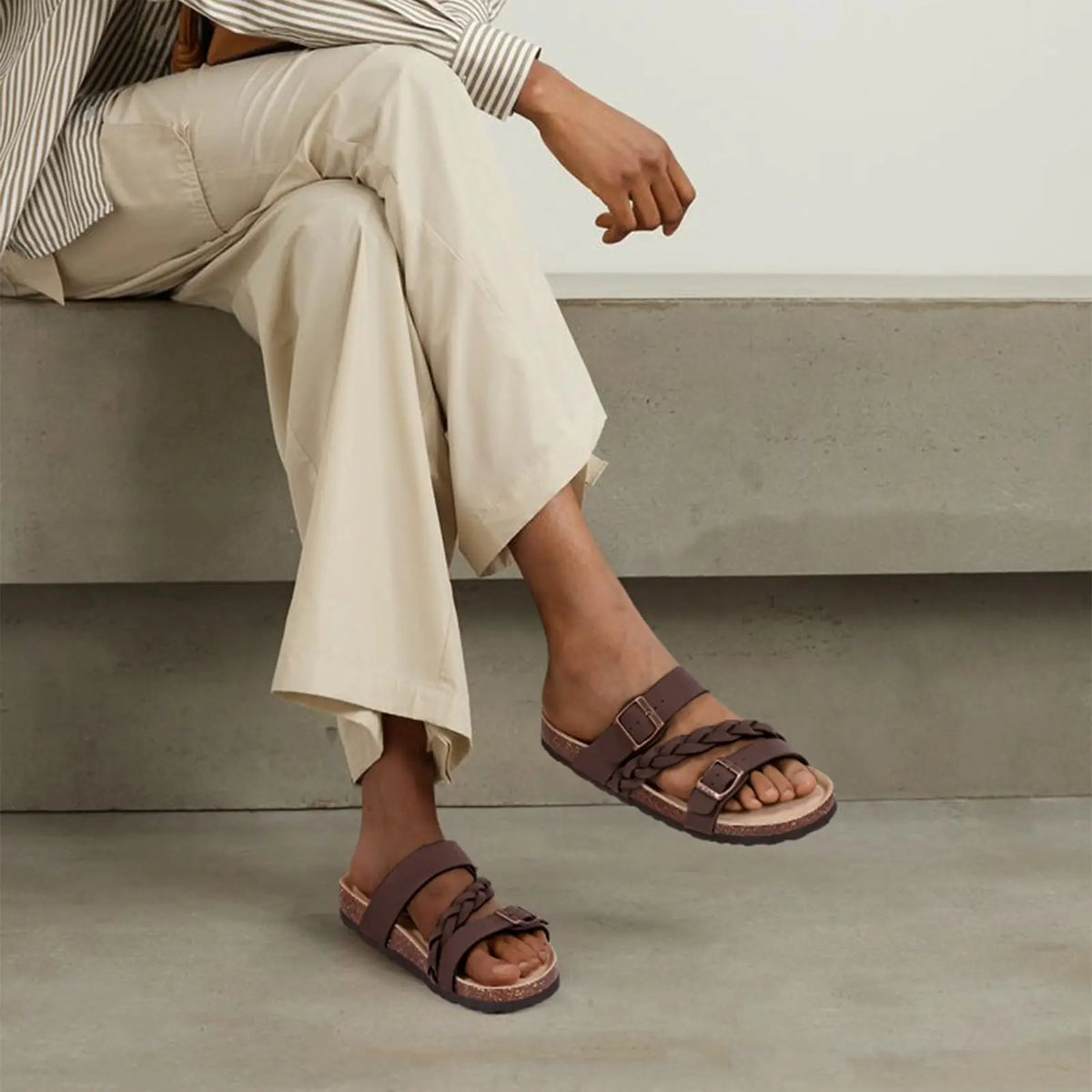 A woman wearing brown cork footbed slide sandals with leather straps, sitting on a concrete ledge.