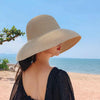A woman wearing an elegant wide brim straw sun hat and a black dress stands by the sea under a clear blue sky.