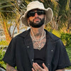 A man wearing a wide-brim bucket hat and sunglasses poses outdoors with tropical foliage in the background.