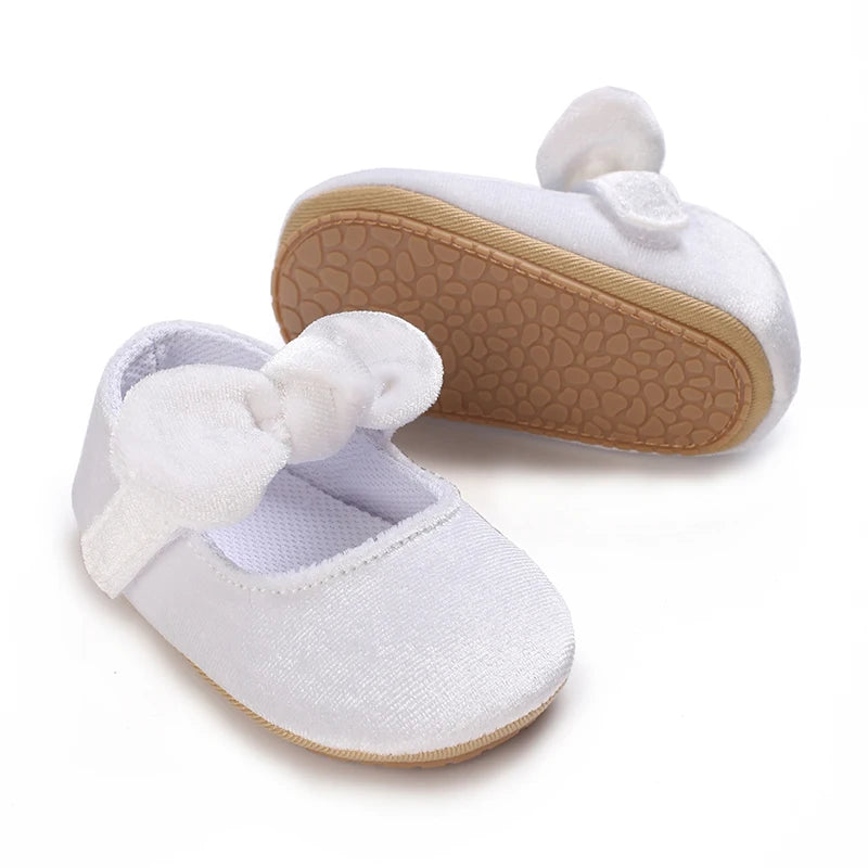 A pair of soft white baby first walker shoes with suede soles and white fabric bows.