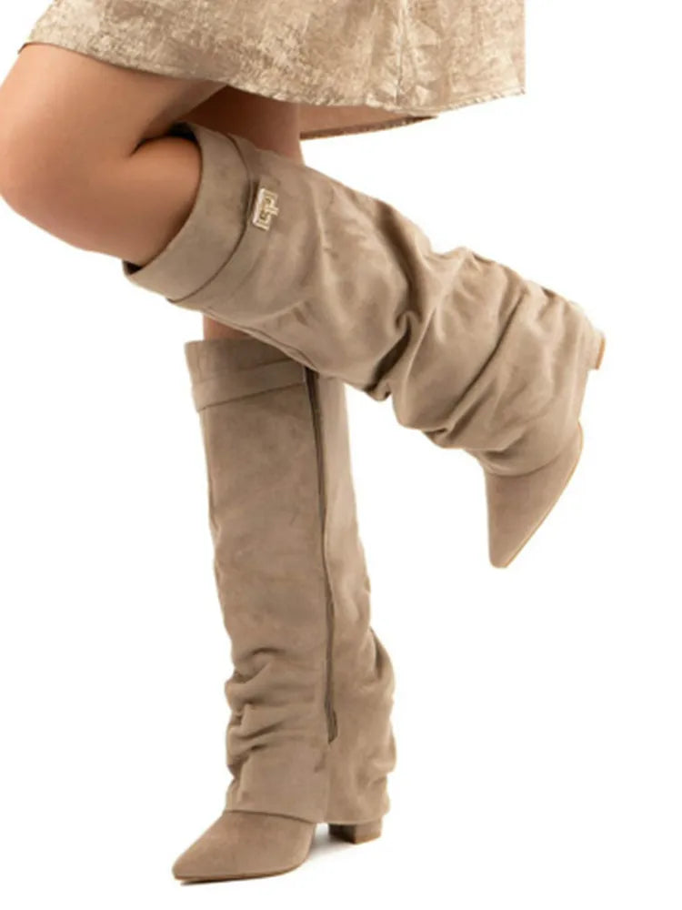 A pair of taupe knee-high chunky heel boots with a metal buckle worn on a foot.