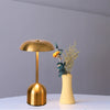 A minimalist gold-toned rechargeable LED table lamp with a dome shade and a vase of dried foliage on a white surface.