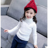 A smiling young girl wearing a white knit turtleneck sweater and a red knitted hat sitting on a gray sofa.