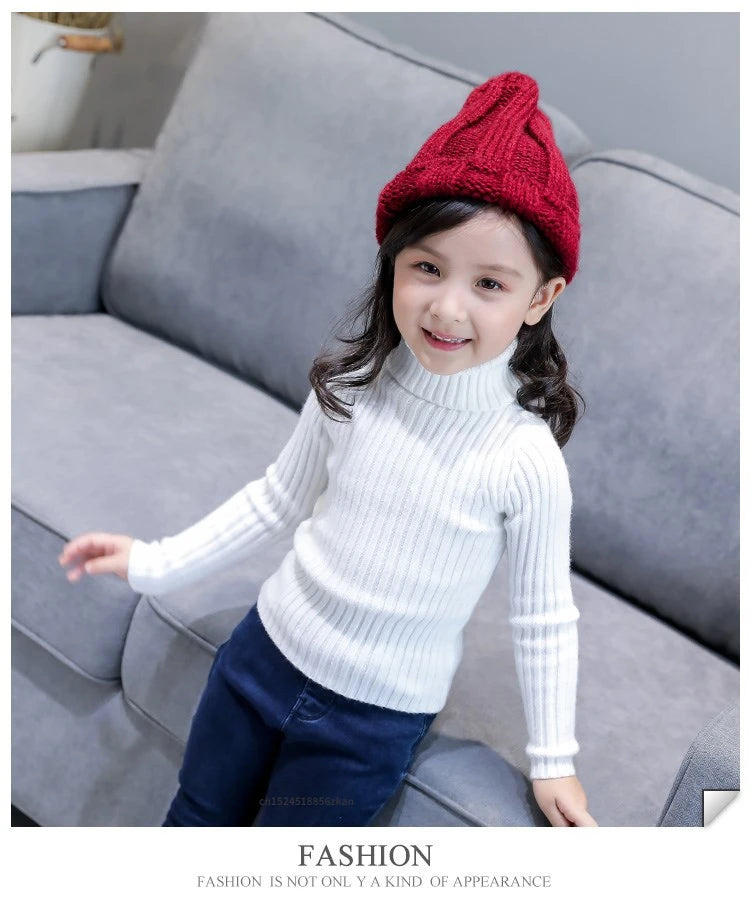 A smiling young girl wearing a white knit turtleneck sweater and a red knitted hat sitting on a gray sofa.