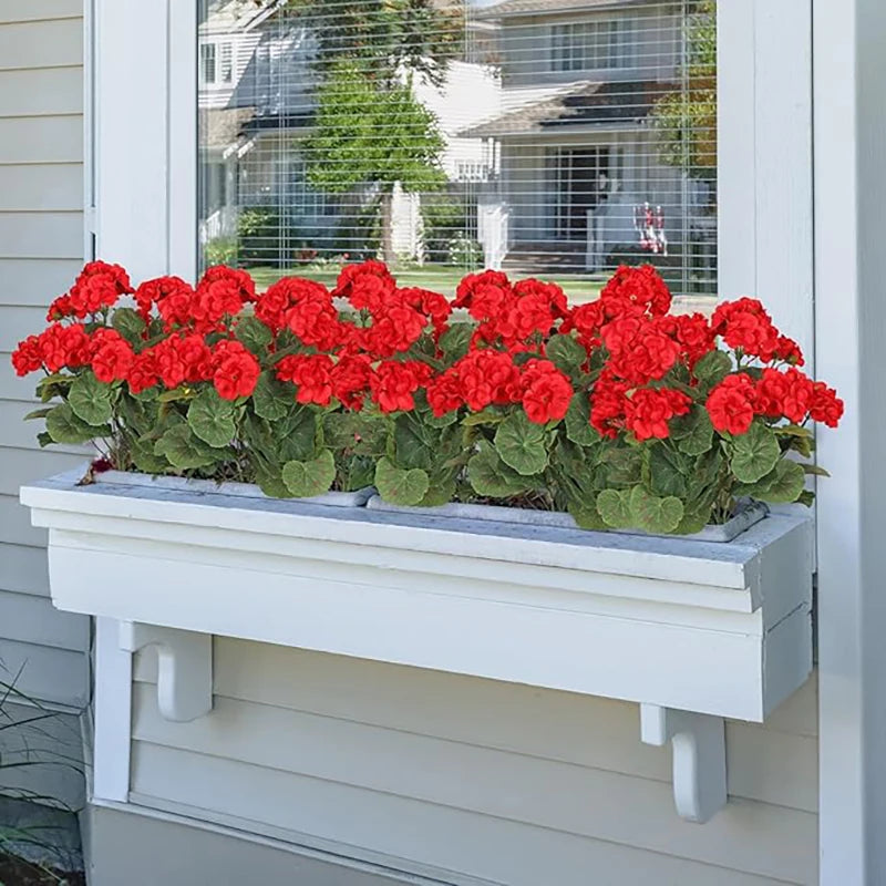 A row of bright red artificial geraniums planted in a white rectangular planter on a light gray siding wall.