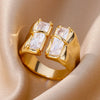 Goldener offener Ring aus Edelstahl mit rechteckigen Zirkon-Akzenten auf beigem Stoff.