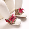 A pair of leopard print baby first walkers with pink laces and soft rubber soles shown on a white sock.