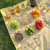 A yellow checkered cloth displays a foldable camping tableware set including bowls, plates, and cups, surrounded by fresh fru