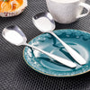 A stainless steel serving spoon rests on a teal plate on a dark textured surface next to a white cup.