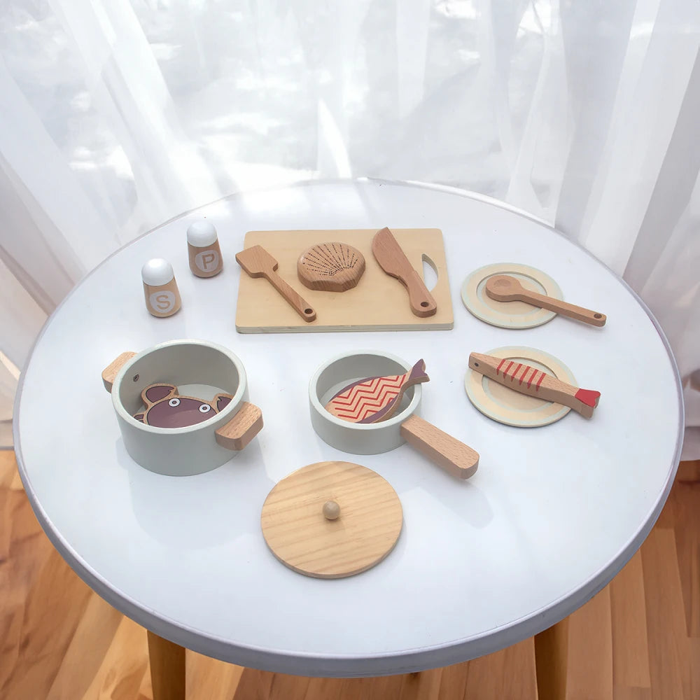 A wooden pretend play kitchen set featuring pots, pans, and play food arranged on a white table.