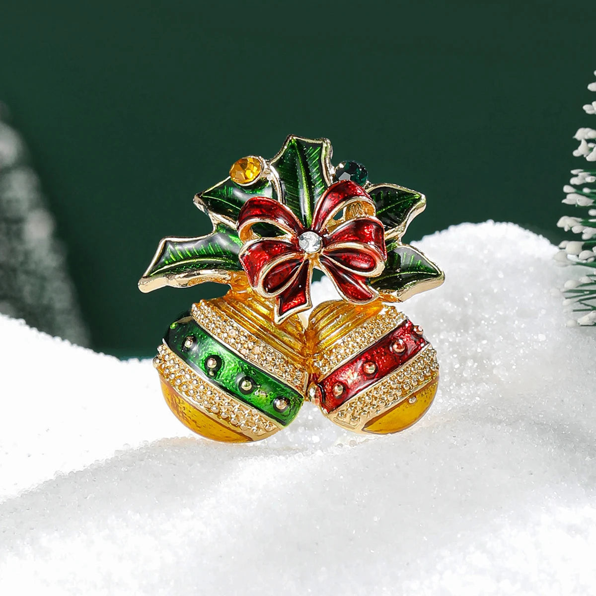 Broche navideño de campana con esmalte y pedrería, acentos rojos y verdes sobre nieve blanca.