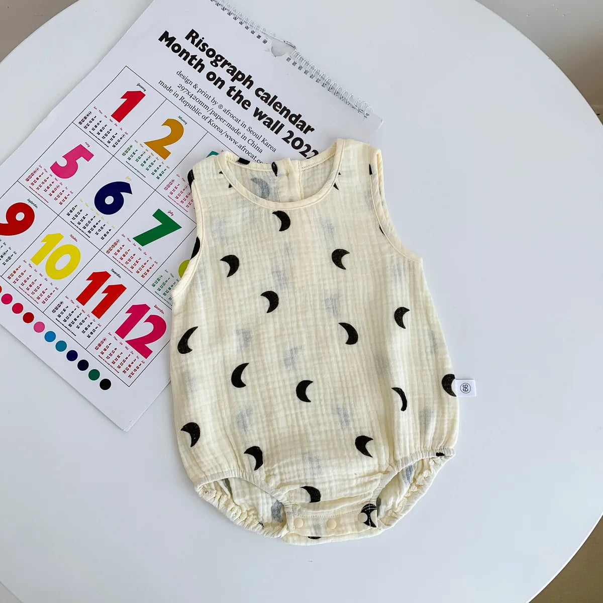 A cream-colored baby cotton sleeveless jumpsuit bodysuit set lying on a white surface next to a calendar.