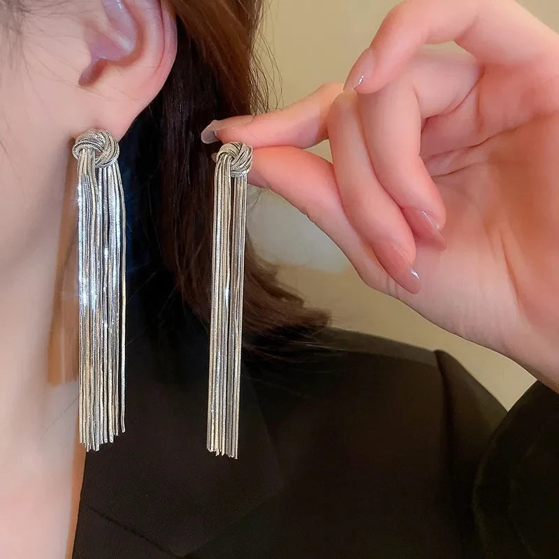 Close-up of a person wearing long, silver geometric tassel drop earrings against a dark background.