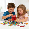 Two children play a bead grabbing game using colored beads and a red bell on a white table.