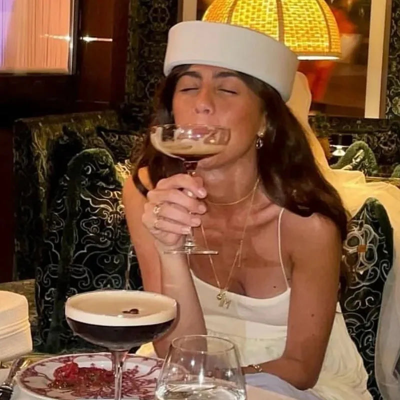 A woman wearing a white straw pillbox sun hat is holding a glass of red wine indoors.