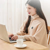 A woman is using a laptop while sitting at a wooden table with a white cup and saucer.