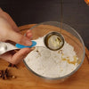 A hand pours oil from a stainless steel measuring spoon into a glass bowl containing white powder and cinnamon sticks.