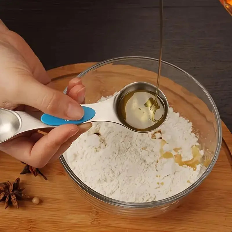 A hand pours oil from a stainless steel measuring spoon into a glass bowl containing white powder and cinnamon sticks.