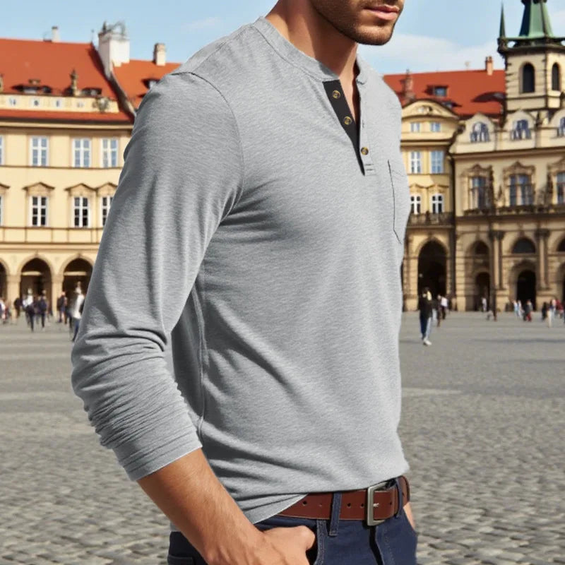 A man wearing a gray long-sleeve shirt with a collar and chest pocket, standing in front of historic European architecture.